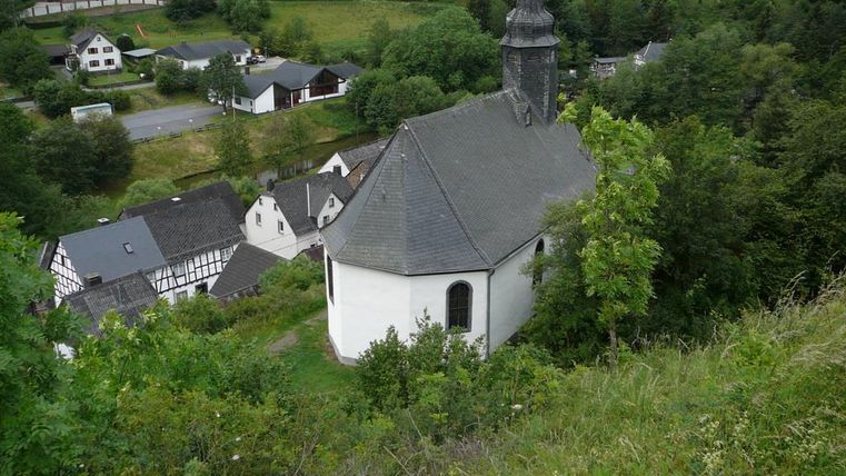 Eine kleine Kirche umgeben von grünen Bäumen und einem malerischen Dorf. Die Gebäude sind traditionell und fügen sich harmonisch in die Landschaft ein.