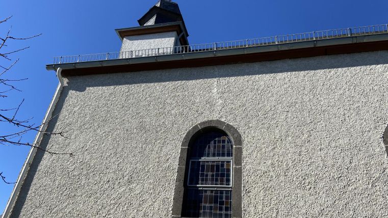 Ein Blick auf die Wand einer Kirche mit einem spitzen Turm und einem blauen Himmel im Hintergrund. Die Fassade ist hell und strukuriert.