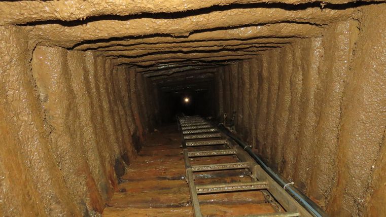 Ein dunkler Tunnel mit einer Holzschiene und unebenen Wänden. Am Ende des Tunnels ist ein schwaches Licht sichtbar.