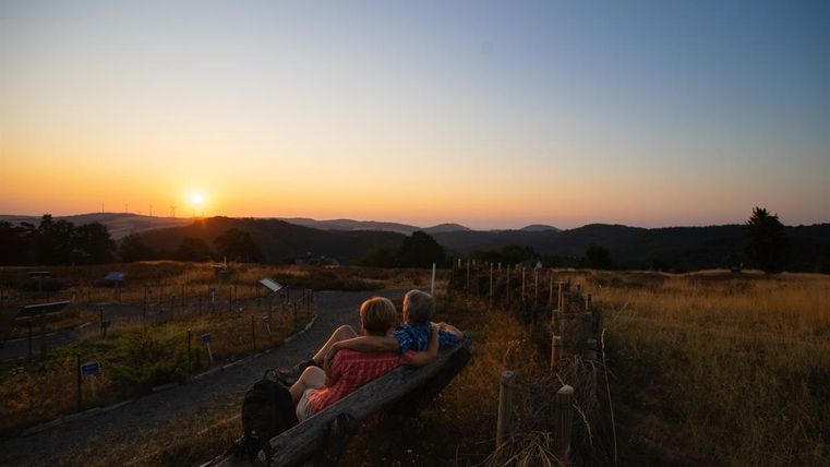 Ein Paar sitzt auf einer Bank und schaut auf den Sonnenuntergang über den Hügeln. Die Landschaft ist von sanften Wiesen und einem klaren Himmel umgeben.