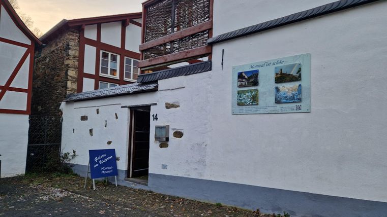 Ein historisches Gebäude mit weißen Wänden und roten Holzrahmen. An der Wand befindet sich ein Informationsschild mit Bildern der Umgebung.