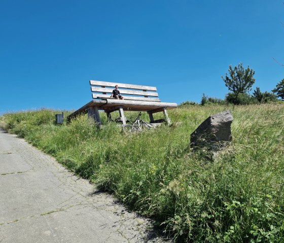 Un &eacute;norme banc en bois est pos&eacute; sur une pelouse verte sous un ciel d&eacute;gag&eacute;. Une personne est assise dessus, &agrave; c&ocirc;t&eacute; d'un v&eacute;lo. Un chemin m&egrave;ne au banc., &copy; Svenja Schulze-Entrup
