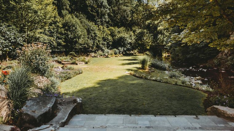 Ein gepflegter Garten mit grünem Rasen und bunten Pflanzen. Im Hintergrund sind Bäume und Sträucher zu sehen.