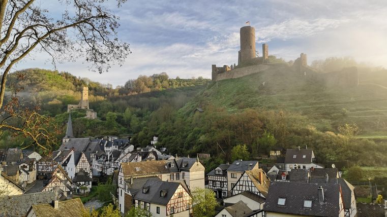 Ein malerisches Dorf mit traditionellen Häusern und sanften Hügeln. Im Hintergrund ragt eine alte Burg in den Himmel.