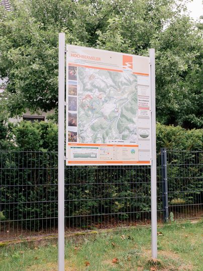 An information sign with a map and various images is set up outdoors. It provides information about the surrounding area and is surrounded by trees.
