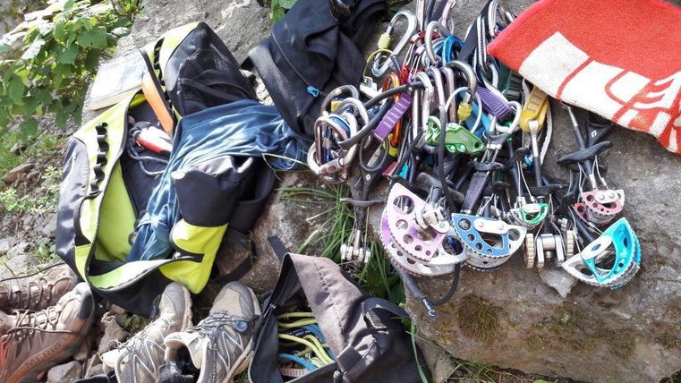 Eine Ansammlung von Kletterausrüstung liegt auf dem Boden, darunter Karabiner und ein Rucksack. Daneben sind Kletterschuhe und ein Handtuch zu sehen.