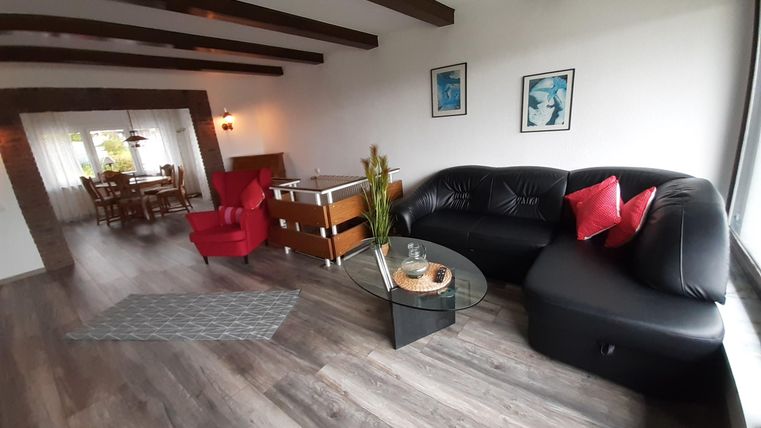 Ein modernes Wohnzimmer mit einem schwarzen Sofa und roten Kissen. Der Raum bietet ein helles Ambiente mit Holzdecken und einem Esstisch im Hintergrund.
