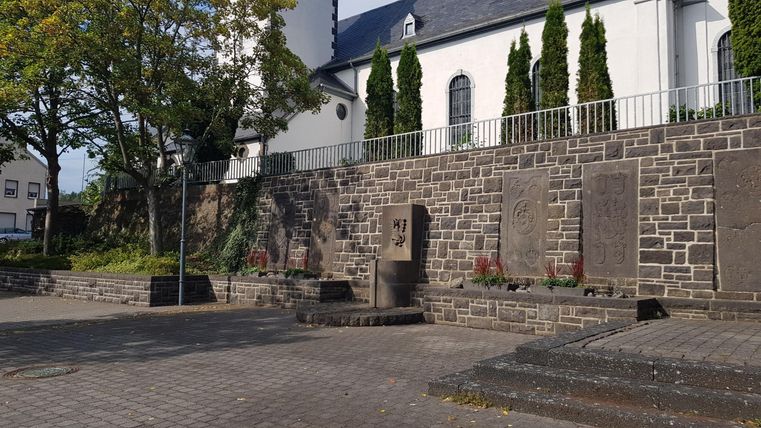 Eine ruhige Stätte mit einer Steinmauer und einem Gebäude im Hintergrund. Grüne Bäume und gepflegte Pflanzen ergänzen die Umgebung.