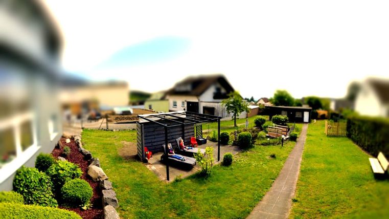 Ein gepflegter Garten mit einer Sitzgelegenheit und grünen Pflanzen. Im Hintergrund ist ein Haus zu sehen.