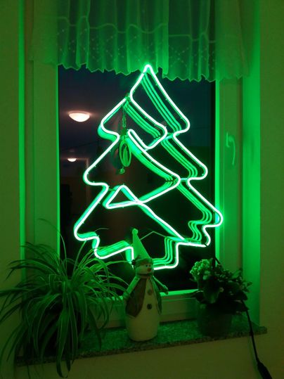 A glowing green Christmas tree made of neon tubes hangs in a window. Next to the tree is a winter decoration with a snowman figure.