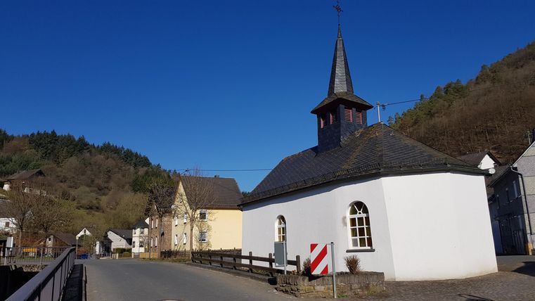 Eine kleine weiße Kirche mit einem spitzen Turm steht an einer ruhigen Straße. Im Hintergrund sind sanfte Hügel und ein klarer blauer Himmel zu sehen.