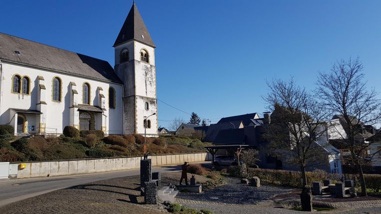 Ein historisches Gebäude mit einem spitzen Turm und einer schönen Fassade. Im Vordergrund ist ein Platz mit Bäumen und einem klaren blauen Himmel.