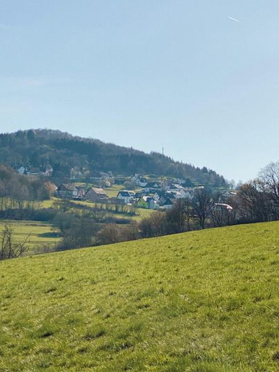Eine ruhige, grüne Landschaft mit sanften Hügeln und einer kleinen Siedlung im Hintergrund. Der Himmel ist klar und blau.