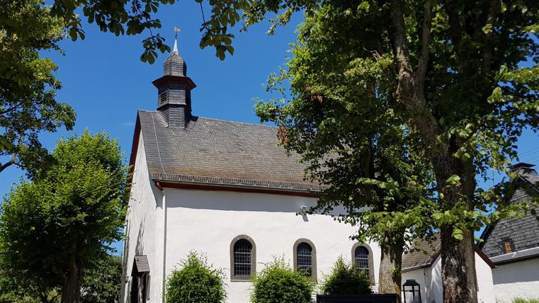 Eine kleine Kirche umgeben von Bäumen und grünem Gras. Der Himmel ist klar und blau.