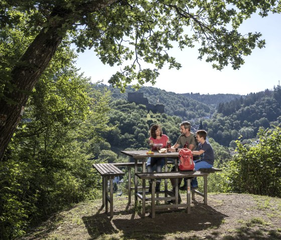 Drei Personen sitzen an einem Picknicktisch im Gr&uuml;nen, umgeben von B&auml;umen, mit Blick auf die Burgruine Virneburg., &copy; Kappest_Remet