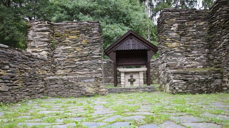 An ancient ruin with stone walls and a small wooden building in the background. Green grass and trees surround the place.