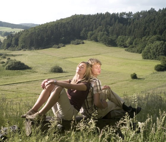 Zwei Personen entspannen auf einer Bank vor einer gr&uuml;nen, h&uuml;geligen Landschaft mit W&auml;ldern im Hintergrund., &copy; Traumpfade