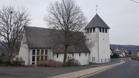 Eine weiße Kirche mit einem Spitzturm steht entlang einer Straße. Im Hintergrund sind kahle Bäume und hügelige Landschaft zu sehen.
