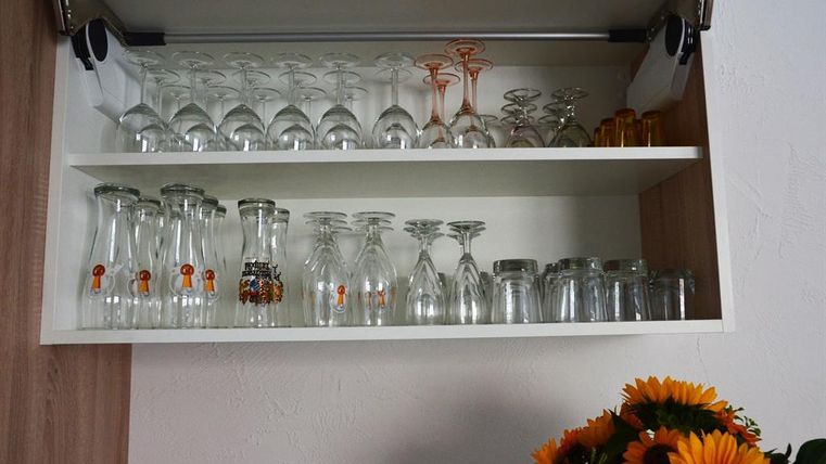 A kitchen cabinet with several shelves, filled with various jars. Below are clear glasses, wine glasses, and decorative bottles.