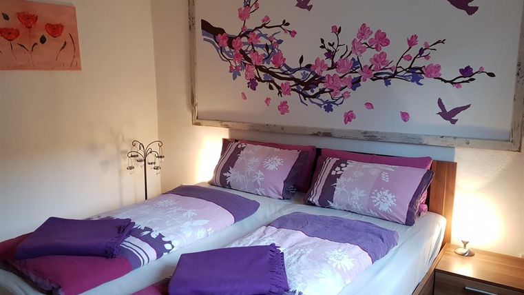 A cozy bedroom with a double bed and purple bed linens. A piece of artwork featuring flowers and birds hangs on the wall.