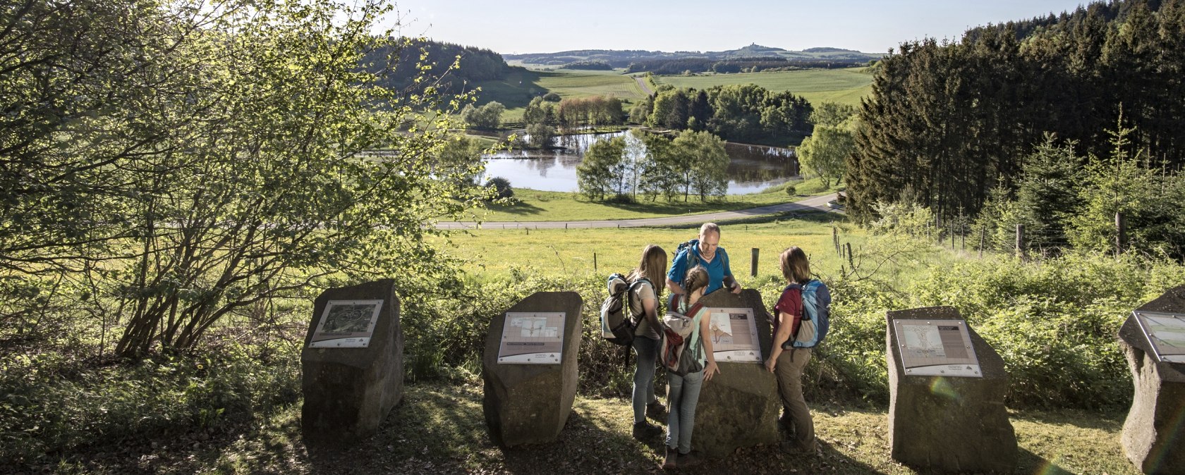 Vier Wanderer betrachten Informationstafeln am Maarkessel Boos. Im Hintergrund eine malerische Landschaft mit See und W&auml;ldern unter blauem Himmel., &copy; REMET, Kappest