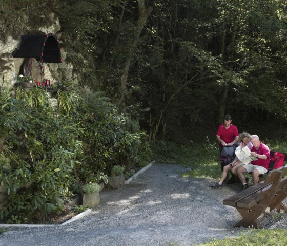 Drei Personen sitzen auf einer Bank im Wald, betrachten eine Karte. Links ist eine Madonna-Statue in einer Felswand zu sehen, umgeben von Pflanzen., &copy; Traumpfade/Kappest