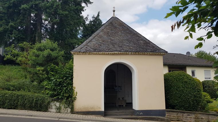 Ein kleiner, runder Pavillon mit einem schiefergedeckten Dach. Umgeben von grünen Bäumen und Sträuchern.