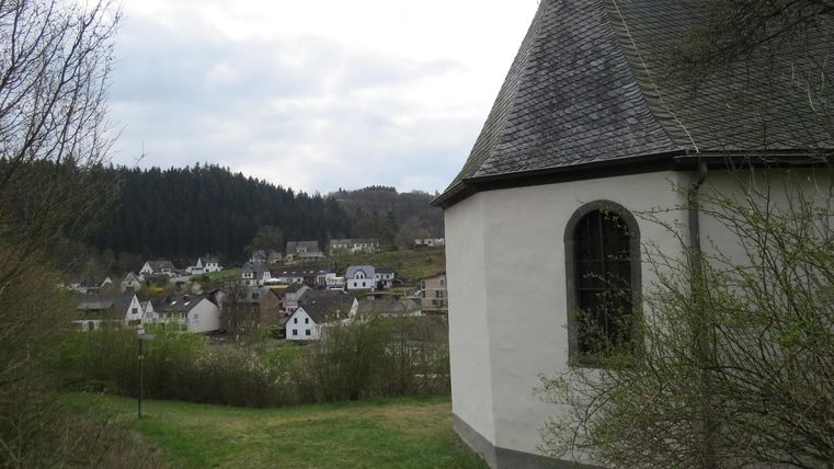 Eine kleine Kirche steht am Hang mit Blick auf ein malerisches Dorf. Im Hintergrund sind Hügel und kühler Himmel zu sehen.