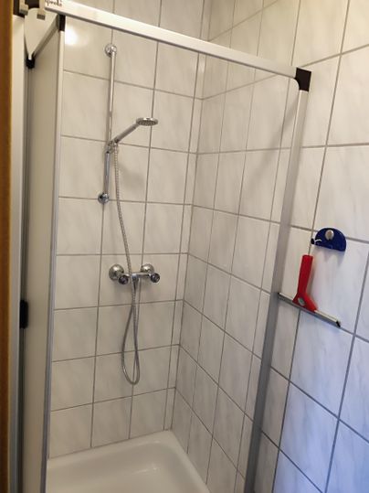 Eine helle Dusche mit weißen Fliesen und einer Glasabschottung. Die Duscharmatur ist an der Wand montiert und es gibt einen Handbrausehalter.