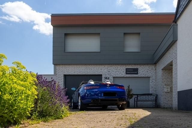 Ein blaues Auto parkt vor einem modernen Haus. Im Vordergrund sind grüne Pflanzen und Blumen sichtbar.