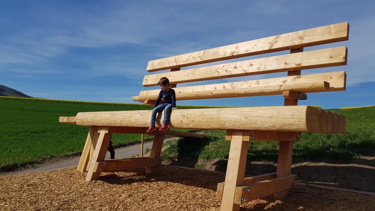 A large wooden bench stands in a green landscape. A child is sitting on the bench and enjoying the view.