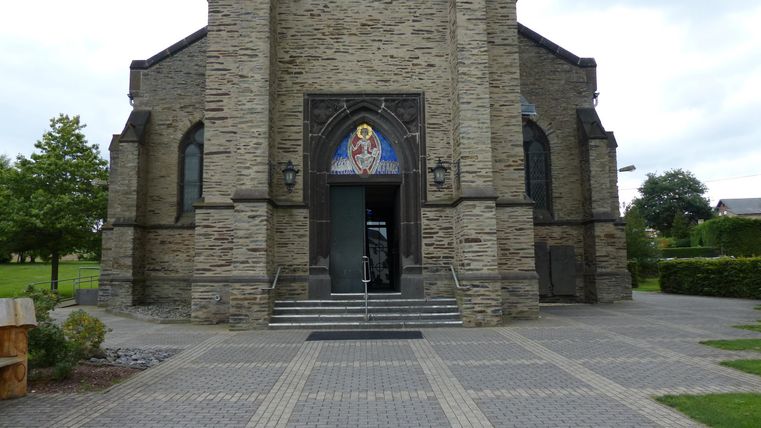 Ein beeindruckendes Kirchengebäude aus Stein mit einer großen Eingangstür. Der Platz vor der Kirche ist gepflastert und von Bäumen umgeben.
