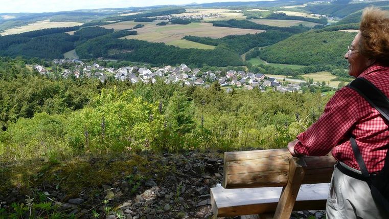A person is sitting on a bench and looking at a picturesque landscape with green hills and a small village in the valley. The sky is blue with some clouds.