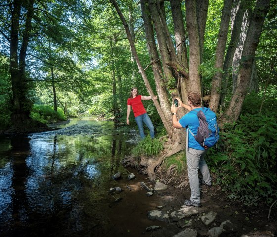 Zwei Personen im Wald, eine posiert auf einem Baum, die andere fotografiert. Ein Bach fließt im Hintergrund, umgeben von üppigem Grün., © kappest_remet