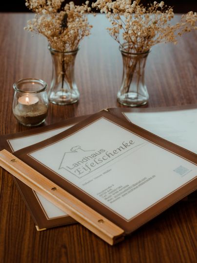 Ein holziger Tisch mit zwei Speisekarten und kleinen Vasen mit Trockenblumen. Eine Kerze sorgt für eine gemütliche Atmosphäre.