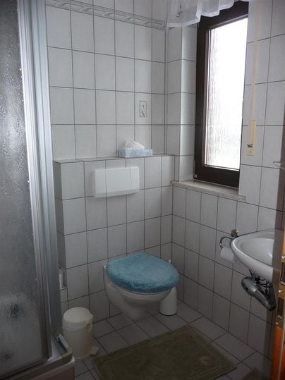 A small bathroom with a shower, a toilet, and a sink. The walls are tiled with white tiles and a window provides daylight.