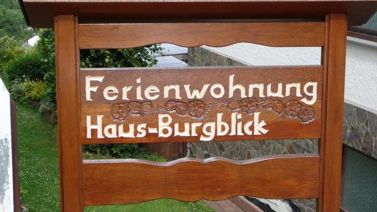 Ein Holzschild mit der Aufschrift "Ferienwohnung Haus-Burgblick". Das Schild ist umgeben von einer grünen Landschaft.