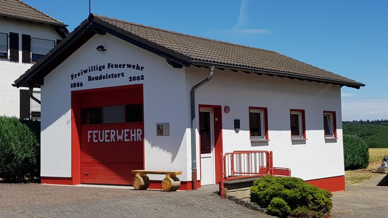 Ein Feuerwehrhaus mit rotem Tor und einer Wetterfahne auf dem Dach. Die Fassade ist weiß mit roten Akzenten und es gibt eine kleine Sitzgelegenheit davor.