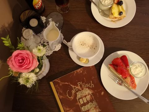 A cozy table with two plates of cake, a coffee cup, and a beautiful flower. In the background lies a menu titled "Café Plüsch."