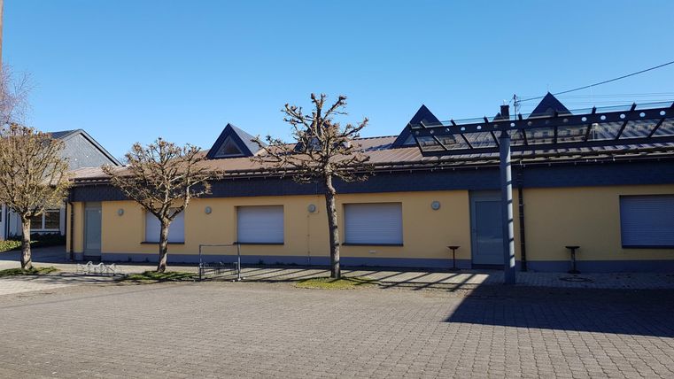 Ein modernes Gebäude mit gelber Fassade und grauen Details. Der Himmel ist klar und blau, und es gibt einige Bäume in der Nähe.