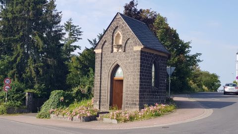 Eine kleine Kapelle aus Stein an einer Straßenkurve. Umgeben von Bäumen und Blumen.