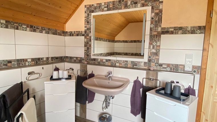 Ein modernes Badezimmer mit Holzdecken und hellen Fliesen. Es verfügt über einen Waschbeckenbereich mit weißen Schränken und handtuchhaltern.