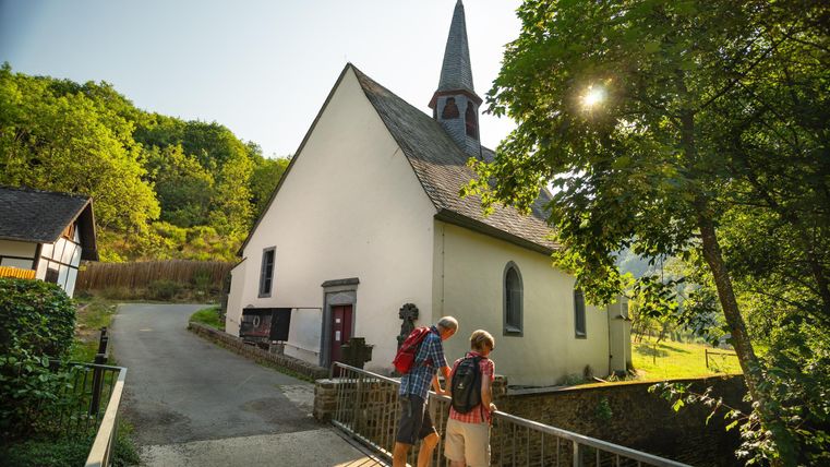 Eine kleine Kirche mit einem steilen Dach und einem hohen Turm. Zwei Wanderer stehen auf einer Brücke und genießen die schöne Landschaft.