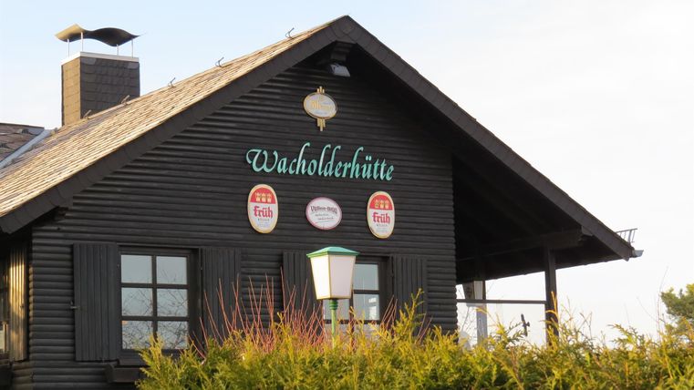 A traditional wooden house with the inscription "Wachholderhütte". It has a dark facade and a small outdoor area with a lantern-like lamp.