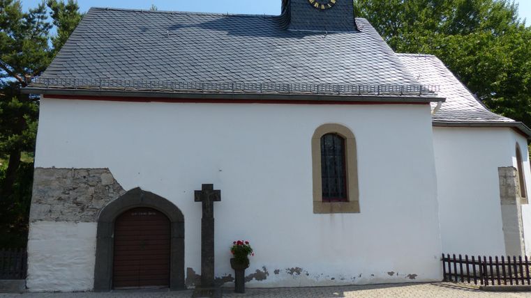 A small white church with a gray roof and a clock on the front. Next to the church stands a cross and flowers are placed at the entrance.
