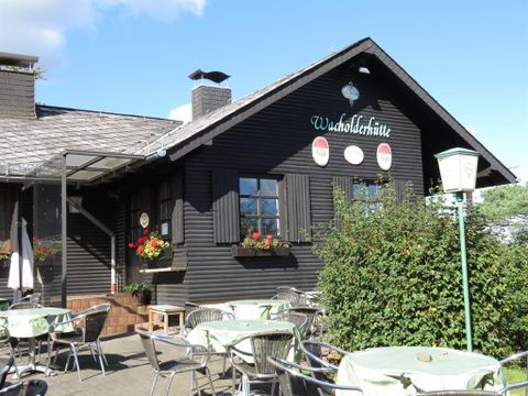 Ein traditionelles, schwarzes Häuschen mit dem Namen "Waldschlötte" steht im Vordergrund. Tische und Stühle stehen im Freien, umgeben von grünen Pflanzen und Blumen.