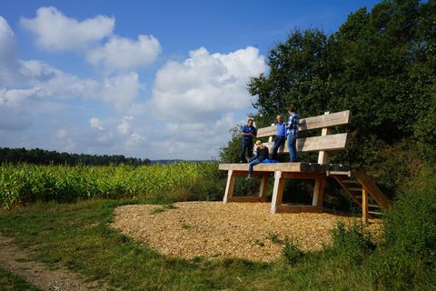 A large wooden bench stands in nature, surrounded by a field and trees. Three children are sitting on the bench, enjoying the view under a blue sky with clouds.