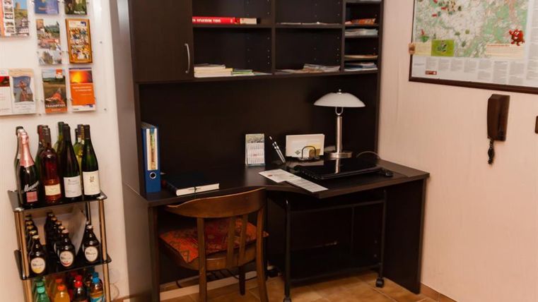 Ein kleines Büro mit einem Schreibtisch, einem Stuhl und einer Lampe. An der Wand sind Karten und Informationsmaterialien sowie ein Regal mit Getränken.