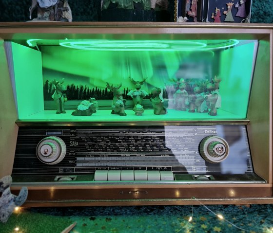 Ein altes Radio mit einer grün beleuchteten Szene von Rentieren und einem Wald im Inneren. Die Beleuchtung erzeugt eine nordlichtartige Atmosphäre., © Svenja Schulze-Entrup
