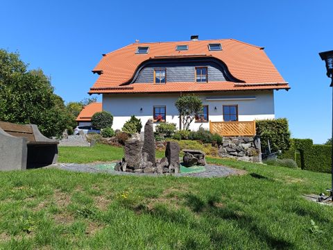 Ein schönes Haus mit einem roten Satteldach und einem gepflegten Garten. Die Umgebung ist grün und einladend, mit einigen Steinskulpturen im Vordergrund.